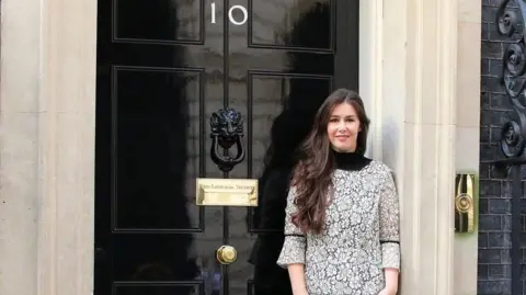Mariposa International Zoe Clark-Coates is standing outside the black door of No.10 Downing Street. She has long dark hair and is smiling. 