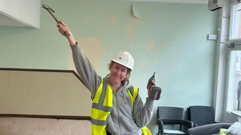 Francesca Griffiths A woman wearing a white hard hat and a green/yellow high-vis waistcoat over a grey hoodie, holds a drill in her left hand and a hammer in her outstretched left hand. She stands in a building with green, peeling paint behind her and chairs stacked behind her.