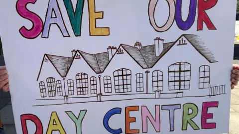 LDRS A Save Our Day Centre sign seen at a protest outside Hove Town Hall over plans to close Wellington House. It has a drawing of a building and a slogan in bright capital letters.