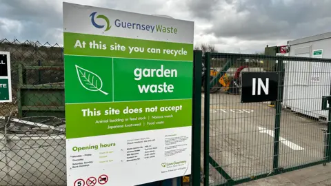 The entrance of a Guernsey Waste recycling centre. There is a large green sign attached to a black gate. It says: At this site you can recycle garden waste. 