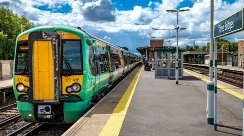 A green and yellow Southern Railway train pulls into Three Bridges Railway Station. It is a sunny day with white, fluffy clouds in the sky.