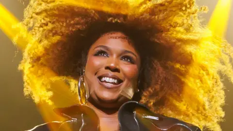 Lizzo on stage in a black leather jacket with two bright yellow lights lighting up her hair