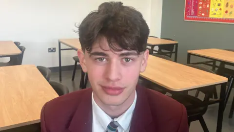 Sean has dark brown hair, short and sides with a fringe that comes just above his eyebrow. His clip on tie is striped and his blazer is maroon. 