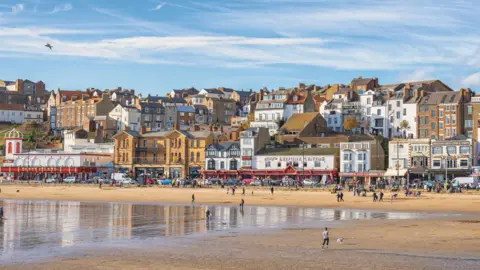 Getty Images A busy seaside promenade with people walking along a sandy beach below a hillside of tightly packed buildings, including fish‑and‑chip shops, an amusement arcade and a leisure centre, on a bright day with a bird flying overhead.