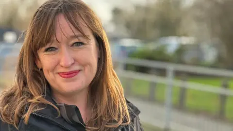 A smiling Emily Cherry has shoulder-length brown hair. She is smiling and wearing a black jacket