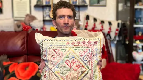 SAM READ/BBC Colin, who has brown curly hair, is holding a multi-coloured embroidered cushion. 