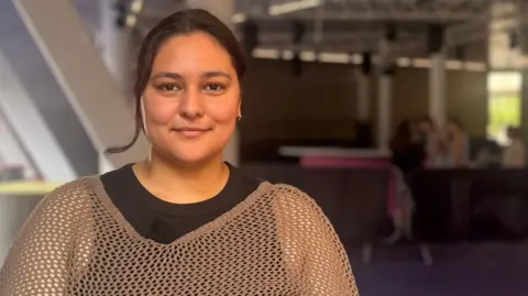 Aaliyah Rice, who is wearing a back top with a knitted top over the top, is smiling at the camera. 