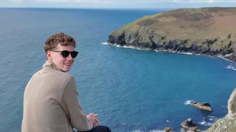 Alfie Watts Alfie is looking over his shoulder sitting on the edge of a hill, with a rocky coastline behind him and a blue sea. He is wearing sunglasses and a light brown sweatshirt. The land arcs into a bay to the right and it is a sunny day.
