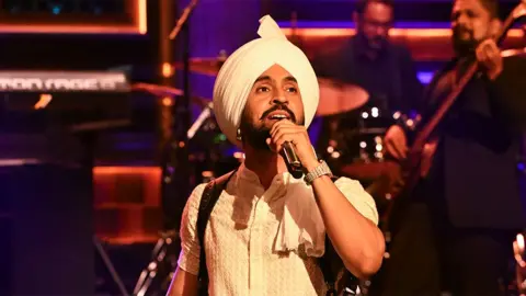 Getty Images Diljit Dosanjh, an artist singing into a black microphone, wearing a cream coloured outfit and turban with a shiny watch on his left wrist. In the background you see two men out of focus playing an instrument.
