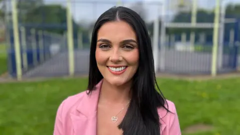 A woman with long dark hair, wearing a pink jacket and a silver heart necklace, standing in a park with a basketball court in the background