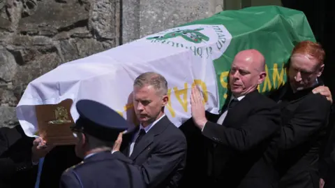 PA Media A number of men in suits carry the coffin of police officer Kevin Flatley which is draped in a green and white GAA flag. A uniformed Garda officer stands to attention in front of the coffin.