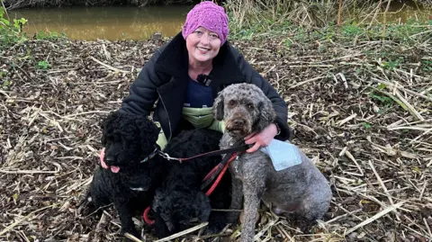 BBC / Naj Modak A woman wearing a pink crochet hat, black fur lined jacket and yellow trousers crouches on the ground with her arm around three medium sized dogs on leads. Two of the dogs are black and the third, on the right, is a grey to brown colour. Behind them is a river.