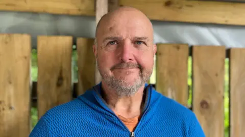 Andy Jones, smiling at the camera. He has a bald head and a short grey beard. He is wearing a blue sweat top.