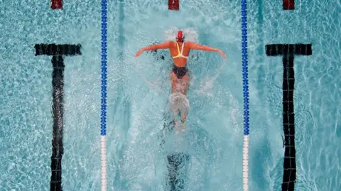 Getty Images A lone woman in a swimsuit and swim cap doing butterfly in the middle lane of a swimming pool with water splashing up from the movement of her arms and legs