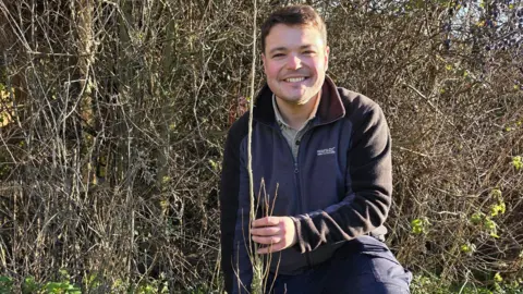 Jake, who wears a grey and black fleece with a beige shirt underneath, smiles at the camera while holding a long thin sapling. There is a hedge behind