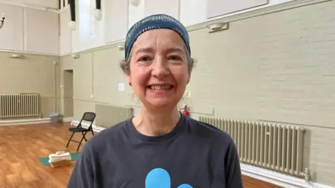 Photo of Massey, who is looking into the camera and smiling. She is wearing a blue headscarf with yellow stripes on it, with her curly brown hair just visible underneath it. Massey is wearing a dark grey t-shirt with a large light blue flower motif just visible. She is stood inside a hall, with has light green and white painted walls and a wooden floor. Behind Massey is a black fold-away chair and a green yoga mat.