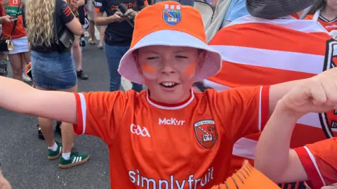 A boy wearing an orange hat an an orange jersey. His arms are stretched out. He's wearing orange face-paint.