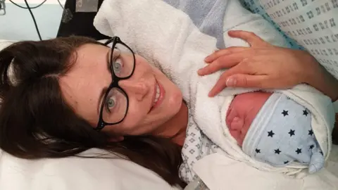 Laura Reed A woman with brown hair and glasses lies in a hospital bed, holding a newborn baby