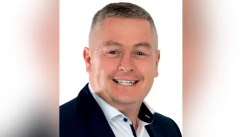 Sinn Féin A close-up image of Councillor Kenneth Collins. He is smiling at the camera in front of a white background. He has short, grey hair and is wearing a navy blazer and white collared shirt.