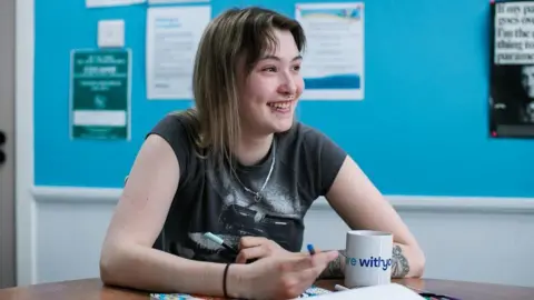 A young woman in a black T-Shirt with long brown hair has a mug in front of her and is smiling
