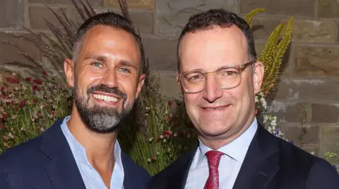 Jens Spahn (R) in glasses, a dark jacket and white shirt and red tie beside his husband Daniel Funke who has as beard and a navy jacket with a blue shirt 