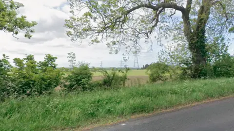 A roadside by Darlington Back Lane in Stockton. It looks over farming land which has brown soil and is surrounded by green shrubbery and trees. There are pylons in the distance. 