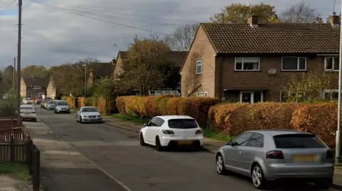 A residential street that is lined with parked cars and houses. There is a bush that runs parallel to one side of the road. The picture has been taken on a grey and cloudy day.