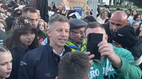 BBC A man in green takes a selfie with a man with short hair and a blue jacket