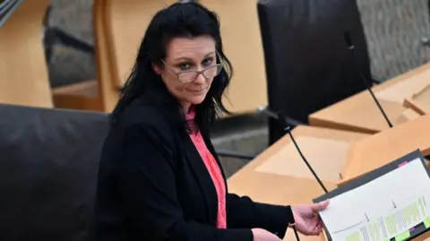 Getty Images Siobhian Brown standing in the Scottish Parliament. She has long black hair and glasses and is wearing a black jacket and a peach top. She has an open folder in front of her but is facing to the side.