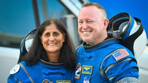 Nasa astronauts Butch Wilmore (on the right) and Suni Williams, wearing bright blue Boeing spacesuits, smile as they pose for pictures before boarding a spacecraft