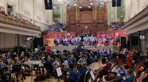 An image of children with a variety of instruments are sitting in different groups on the lower floor of a large building. On the stage in the background sits an orchestra made up of adults.