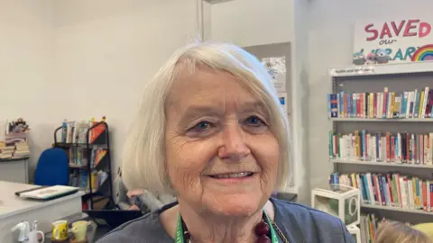 Laura Coffey/BBC A woman with white hair wearing a dark blue jacket, a green lanyard and a red bead necklace. There are books in the background. She is smiling at the camera.
