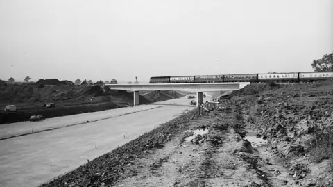Cumbria Railways Association A black and white image of a train going over the old Clifton bridge, while the M6 motorway is being constructed underneath it. There is a smooth layer of concrete that will become the road. The inclines either side are dug up, with piles of mud dotted around.