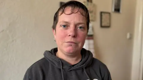 Nicola Cooper has short dark hair and blue eyes. She has an eyebrow and lower lip piercing and is wearing a dark grey/brown hoodie. She is standing in a room with cream walls and some blurred out pictures on the wall on the right.