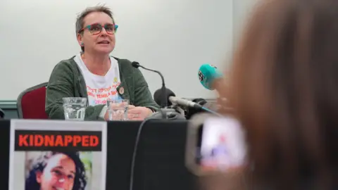 PA Media Mandy Damari, a woman with short blonde hair wearing a white t-shirt and green cardigan, sits behind a desk talking into a microphone. On the desk is a poster with the title 'kidnapped' and a photo of Emily Damari.