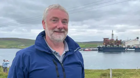 Deputy Chairman of Lerwick Port Authority, Colin Nicolson wearing a blue waterproof jacket stands on a grassy shoreline overlooking a harbor. In the background, the Allseas heavy-lift vessel Pioneering Spirit carries a large offshore platform topside, with cranes visible on deck. The scene is set under an overcast sky, with low hills and water stretching across the horizon.