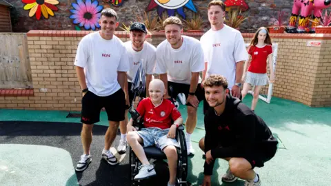 Bristol City FC Five Bristol City players, four of them wearing white T-shirts and shorts, pose with a young boy who is sitting in a wheelchair wearing a red top and light blue denim shorts