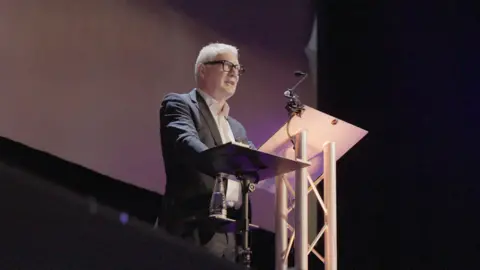 WMCA West Midlands mayor Richard Parker, a man with short white hair and black-rimmed glasses, stands at a lectern with a microphone in front of him. He is wearing an open-necked white shirt under a navy suit jacket.