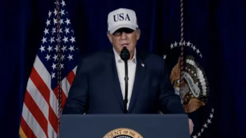 US President Donald Trump stands at a podium wearing a white cap with 'USA' written on it