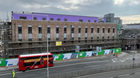 A picture of building works taking place in Plymouth. There is a brown building with a road in front of it. There is a red double decker bus.