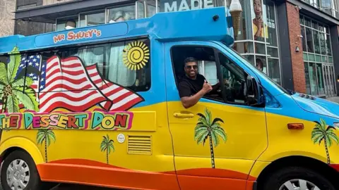Mr Shuey Mr Shuey in his ice cream van which has palm trees, the US flag, and yello, blue and orange wave designs.