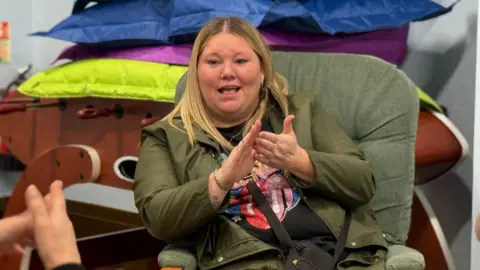 Charlie O'Loughlin A woman with blonde hair signs with her hands as looks off camera towards a person not in shot. She wears a green jacket, a colourful T-shirt and has a black bag across her body. She is sitting on a high-backed green armchair, and behind her is a table football game with a green, purple, and blue bean bag stacked on top. 