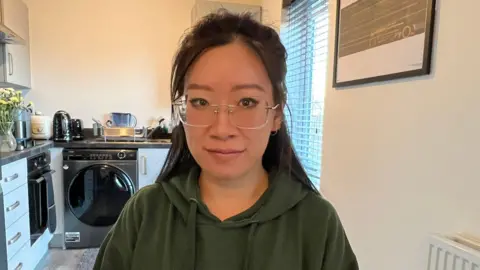 The BBC looks at the camera with a neutral expression in a kitchen. She wears frimless glasses and a green hoodie and her black hair hangs behind her shoulders. There is a washing machine on the left side of it and a vase of yellow flowers is on a worktop next to it.