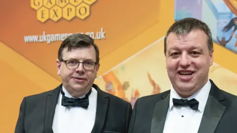 Games Expo Two white men with dark, short hair, wearing tuxedo suits, black dicky bows and a white dress shirt. They are stood in front of a yellow sign that reads Games Expo. 