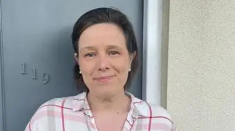 Plymouth City Council Carol Ney wearing a pink, red and white check shirt and standing in front of a grey door