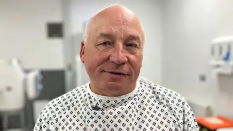 Peter Moult looks into the camera while wearing a hospital gown at Coalville Community Hospital