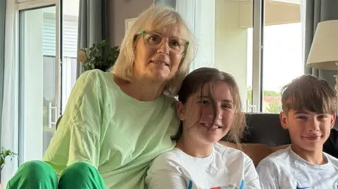 Miriam Norrish Miriam sitting on a sofa beside her teenage granddaughter and grandson, all smiling to camera