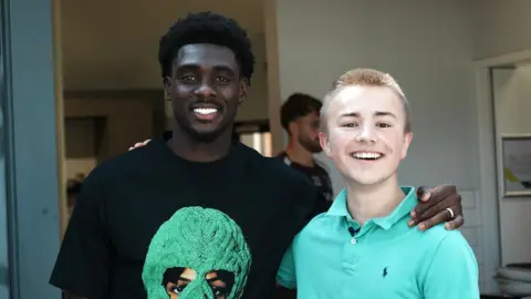 Norwich striker Mo Touré (left) is wearing a black T-shirt with a large green design on the front. He is standing in an office next to 16-year-old fan Ranulph Bacon, who is wearing a green polo shift. The pair are smiling at the camera.