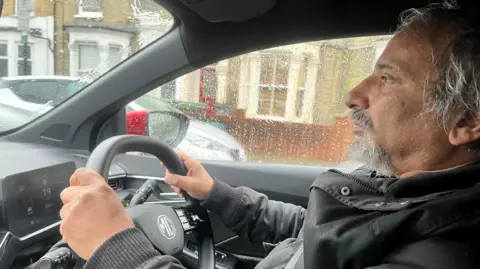 John Sinha in a black waterproof coat sat inside a car. His hands are on the steering wheel and he is looking out of the front windscreen. 