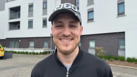 Jose Alves is wearing a black jacket, white t-shirt and grey hat 